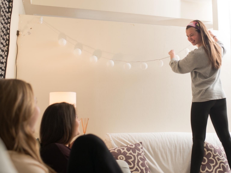 female student hanging fairy lights along dorm room wall 